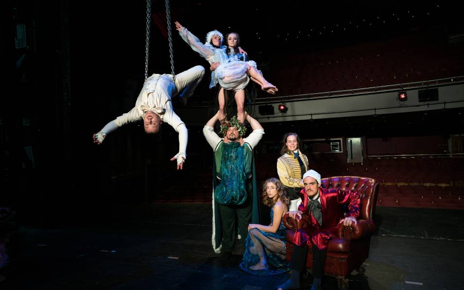 An acrobat hanging backwards off chains in the air. A strongman carrying two other performers on his shoulders and other members of Lost in Translation Circus around them