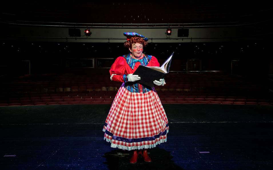 Richard Gauntlett dressed as a dame, holding a large storybook, on stage at the Theatre Royal